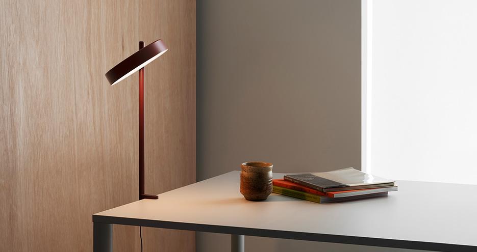 Schreibtisch mit einer dunkelroten Tischlampe (Pastille von Wastberg+) links, einem kleinen braunen Keramikbecher und einem Stapel Bücher auf einer hellen Tischplatte; im Hintergrund eine helle Wand mit Holzpaneel und seitlichem Lichteinfall.