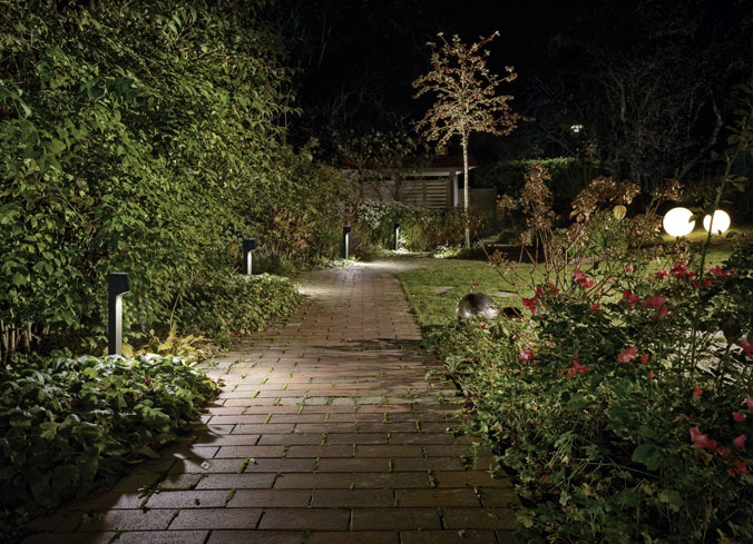 Ein Foto von einem Garten bei Nacht, der gepflasterte Weg wird von Pollerleuchten erhellt.