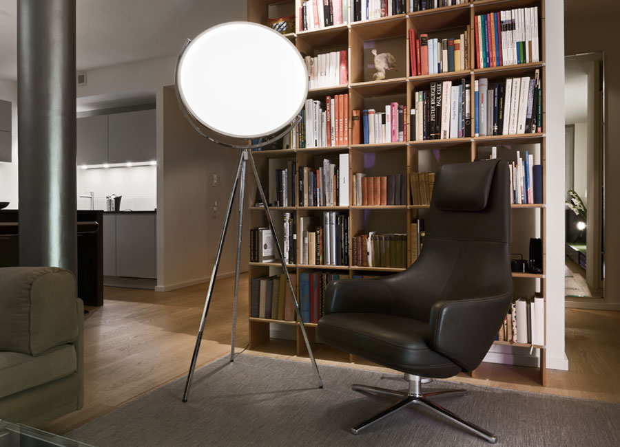Vor einem Bücherregal steht ein Sessel, daneben die Standleuchte Superloon mit ihrer großen, kreisrunden Scheibe als Lichtquelle. (Foto: Prediger Lichtberater)