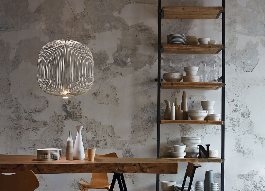Eine Spokes 2 Midi Pendelleuchte hängt uber einem schlichten Holztisch, rechts im Hintergrund ist ein Regal mit Vasen und Tellern zu sehen. (Foto: Foscarini)