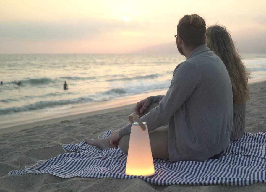 Ein Pärchen sitzt mit einer Uma Mini Soundlaterne bei Sonnenuntergang am Strand. (Foto: Pablo Designs)
