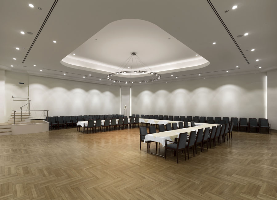 Ein großer Konferenzraum mit Downlights in Wandnähe, einem Kronleuchter sowie Voutenbeleuchtung an der Decke und in den Ecken. (Foto: Prediger Lichtberater)