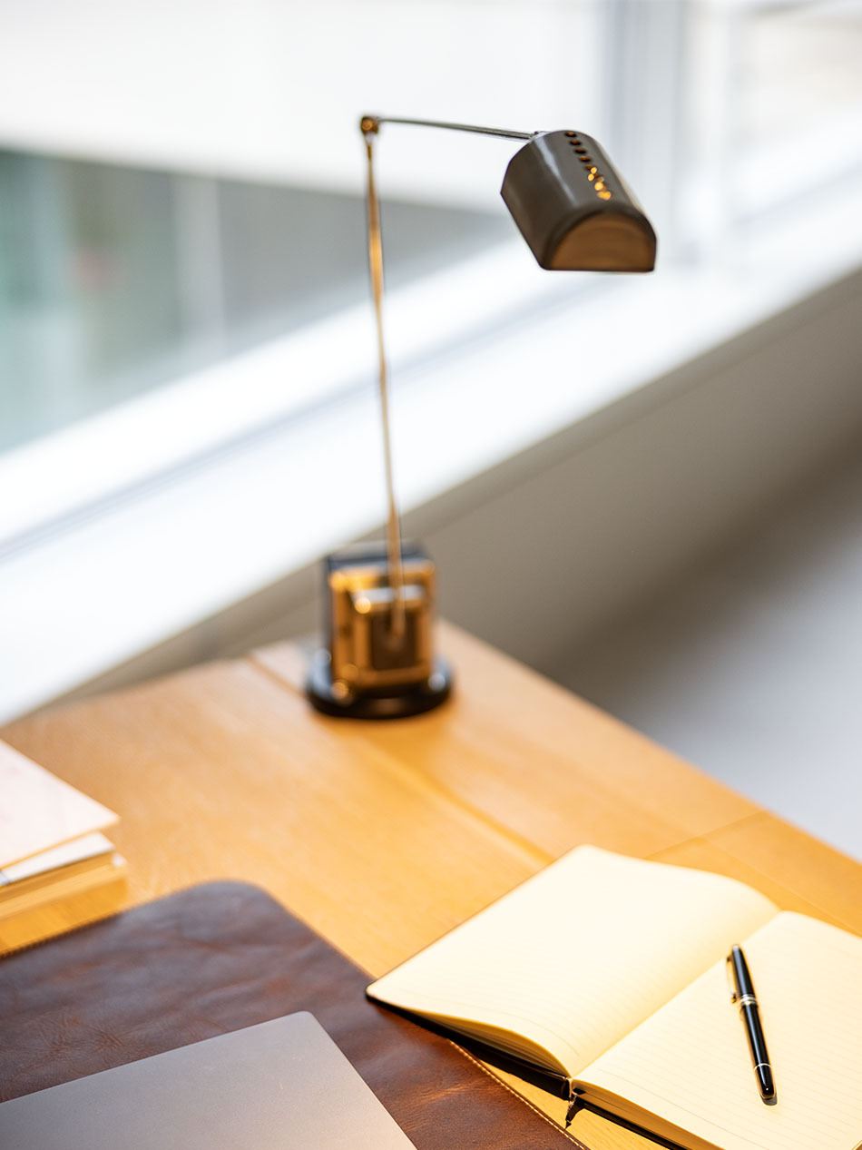Arbeitszimmer mit einer Daphine Tischleuchte von Lumina in Gold. Sie beleuchtet einen Schreibtisch aus Holz, auf dem ein Notizbuch und ein Laptop liegt.