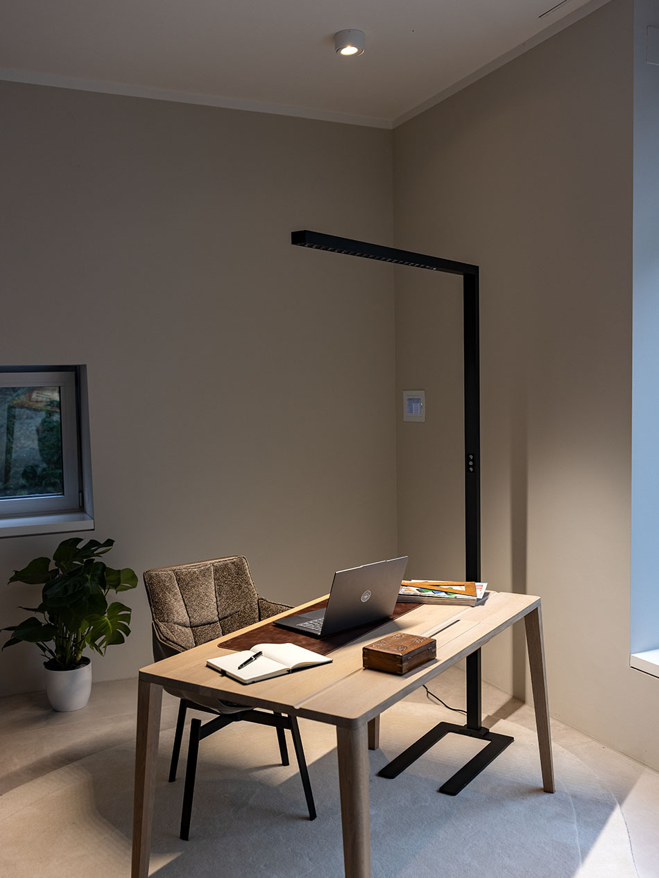 Modernes Arbeitszimmer mit p.110 Office-Standleuchte von prediger.base, die neben einem Schreibtisch steht. Auf dem Schreibtisch befindet sich ein Laptop, ein Notizbuch sowie mehrere Magazine.