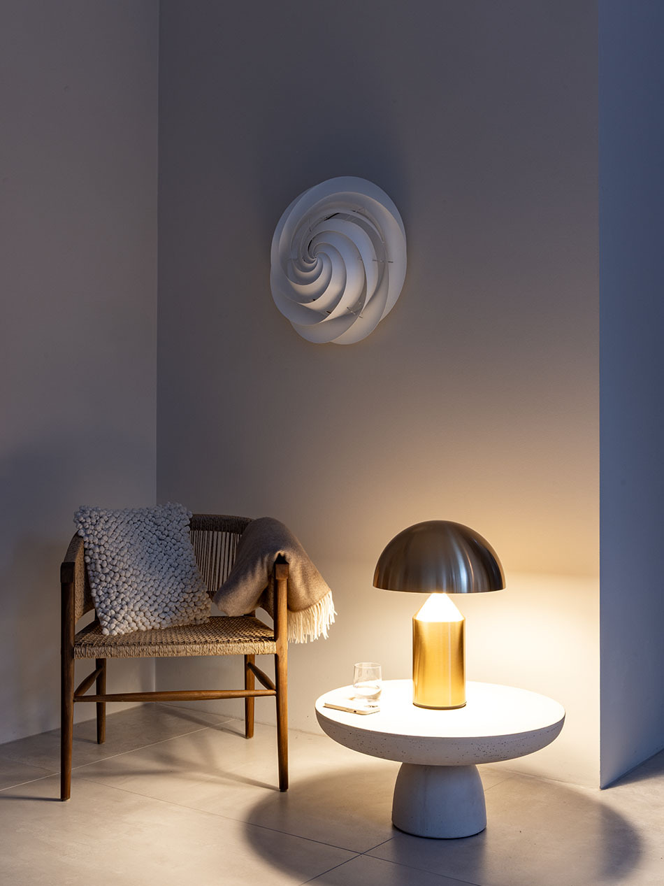 Das Bild zeigt eine gemütliche, minimalistische Wohnszene. Ein Holzstuhl mit geflochtenem Sitz und Decke steht neben einem runden Beistelltisch. Auf dem Tisch befindet sich die goldene Tischleuchte Atollo von der Marke Oluce, die warmes Licht ausstrahlt.