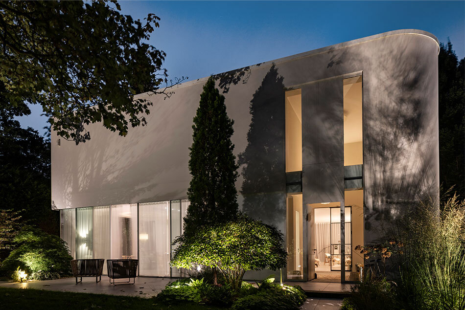 Das Bild zeigt ein Haus in nächtlicher Dunkelheit, das von innen warm beleuchtet ist. Strahler erhellen den Garten, und auch der Weg ist beleuchtet.