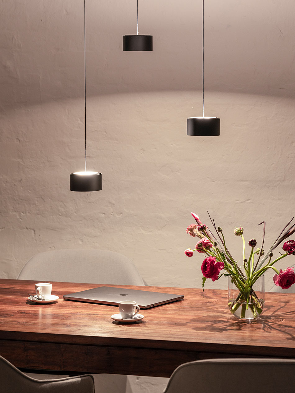 Über einen Holzesstisch hängen drei schwarze s.200 Pendelleuchten von prediger.select auf unterschiedlichen Höhen. Auf dem Esstisch befindet sich ein Macbook, zwei Esspresso-Tassen und ein Blumenstrauß.