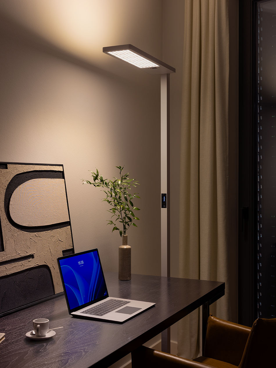 Eine p.145 Office-Standleuchte von prediger.base in Pepper White beleuchtet einen schwarzen Schreibtisch mit Laptop und Blumenstrauß. Die Leuchte hat einen integrierten Display zum Bedienen der Leuchte.