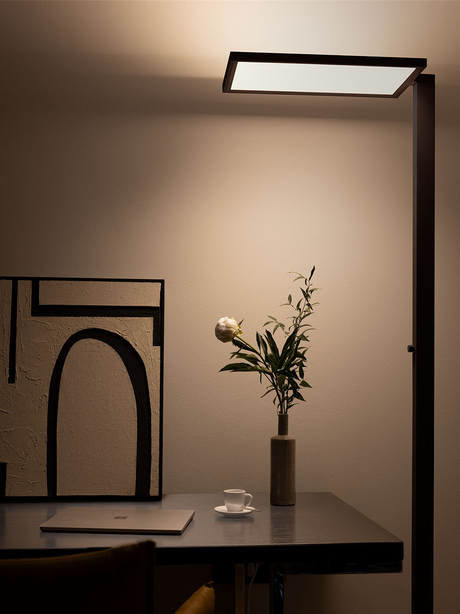 Minimalistisches Büro mit p.144 Office-Standleuchte von prediger.base in Earth Black . Die Stehleuchte beleuchtet einen schwarzen Schreibtisch, auf dem ein Laptop, eine Kaffeetasse und ein kleiner Blumenstrauß steht.