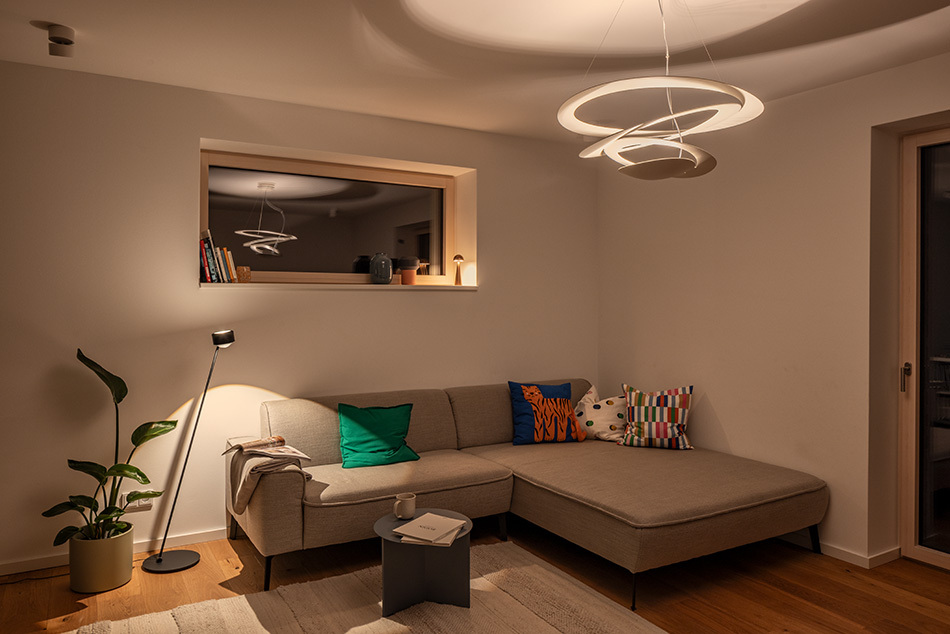 Modernes Wohnzimmer mit einer weißen Pirce Pendelleuchte von Artemide. Neben dem grauen Ecksofa steht eine s.200 Standleuchte von prediger.select in Satin schwarz. Auf der Fensterbank steht die p.141 Akkuleuchte von prediger.base. Außerdem befindet sich an der Decke ein p.011 Deckenstrahler von prediger.base.