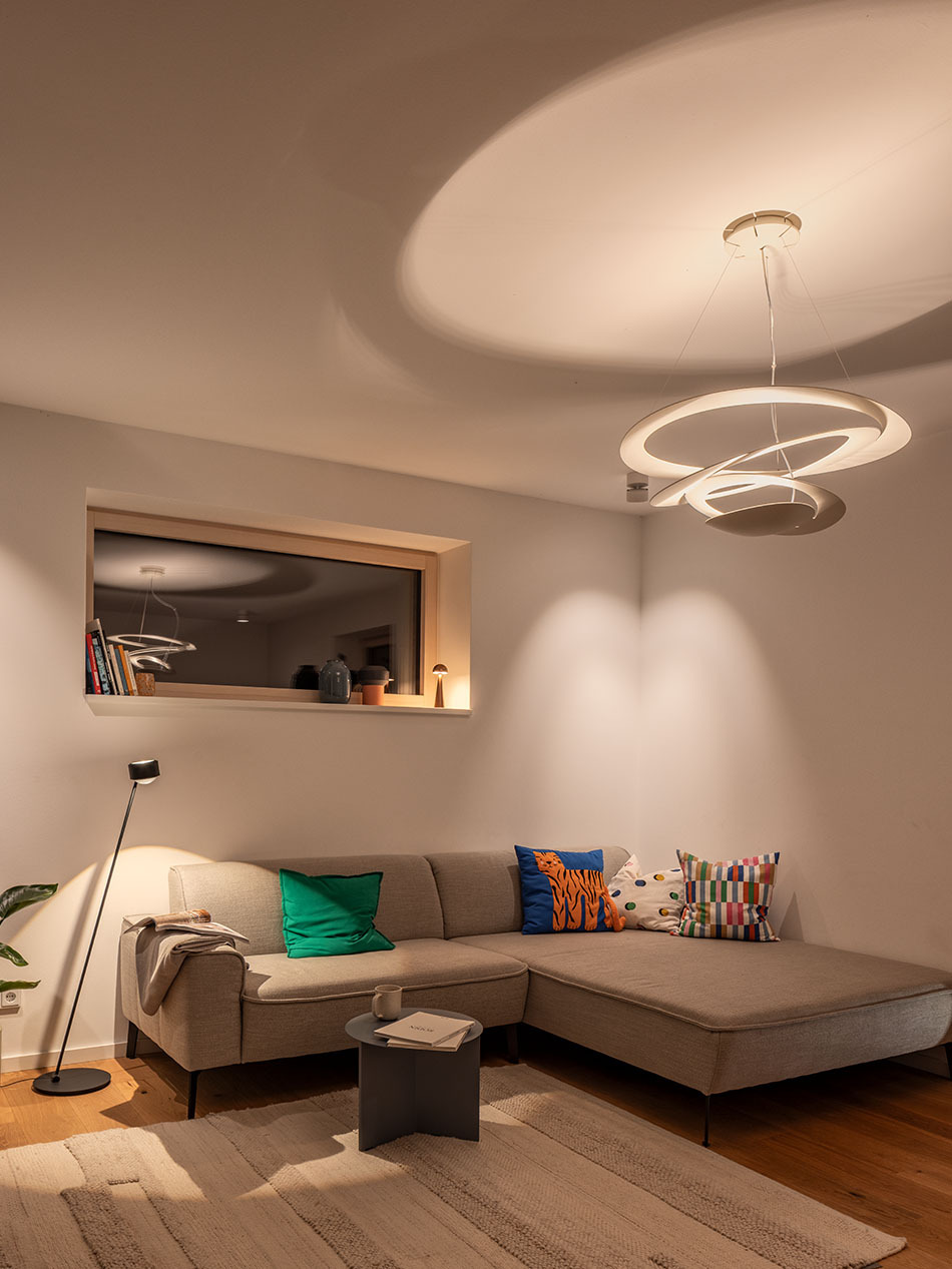 Wohnzimmer mit einem grauen Ecksofa und einer s.200 Standleuchte von prediger.select. Auf der Fensterbank steht eine p.141 Akku-Tischleuchte von prediger.base. Der Raum wird von einer Pirce Pendelleuchte von Artemide beleuchtet.