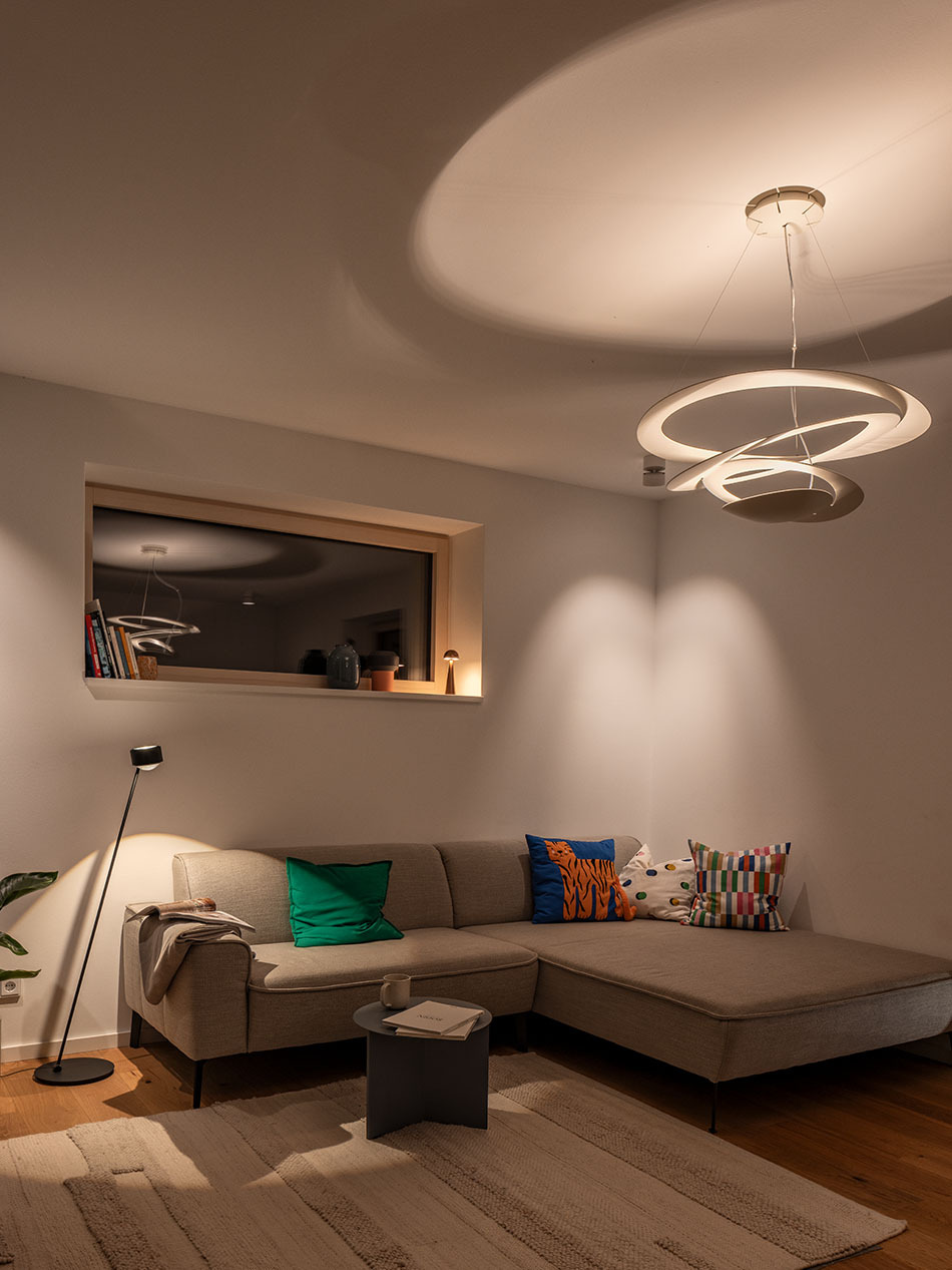 Wohnzimmer in einem Neubauhaus mit der Pirce Pendelleuchte von Artemide in weiß. Neben dem grauen Sofa steht eine s.200 Standleuchte von prediger.select in satin schwarz. Auf der Fensterbank steht eine p.141 Tisch- und Akkuleuchte von prediger.base.