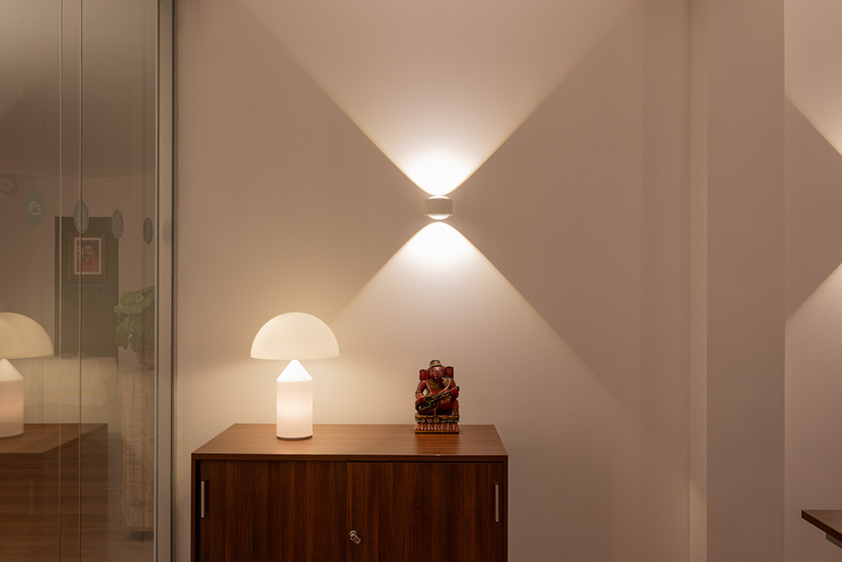 Auf einer Holzkommode im Arbeitszimmer steht eine weiße Atollo Tischleuchte von Oluce. Über der Kommode wurde eine weiße p.133 Wandleuchte von prediger.base montiert, die kegelförmiges Licht nach oben und unten abgibt.