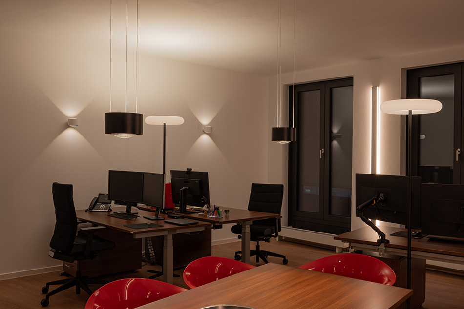 Das Büro wird von zwei p.133 Wandleuchten von prediger.base beleuchtet. Vertikale LED-Wandprofile von Sigor sowie zwei Oto Standleuchten von Belux sorgen für die Allgemeinbeleuchtung. Über einen Konferenztisch hängen zwei schwarze Pendelleuchten von prediger.base.