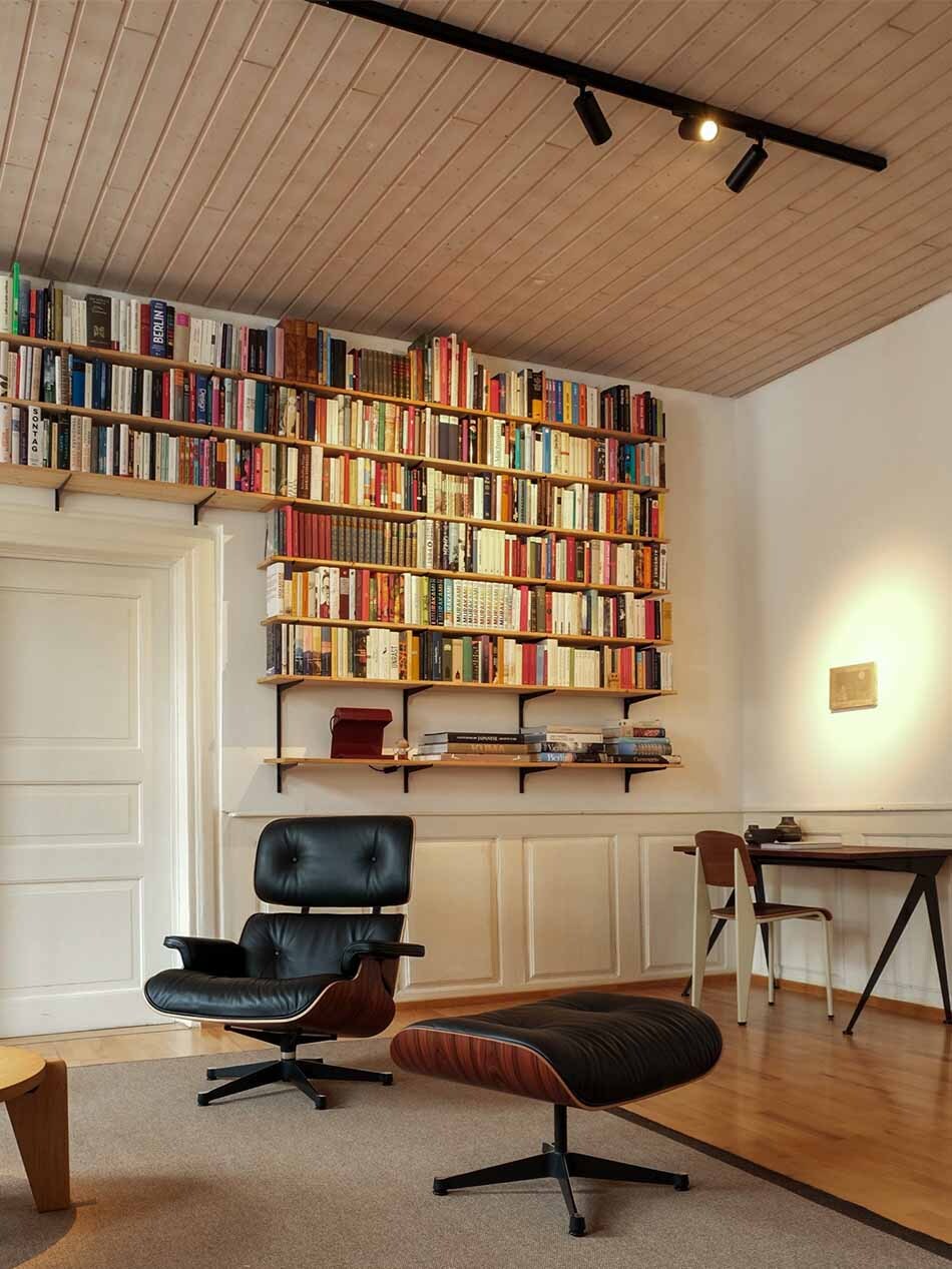 Wohnzimmer mit einem großen Bücherregal, das von p.083 Schienenstrahler in schwarz von prediger.base beleuchtet wird.