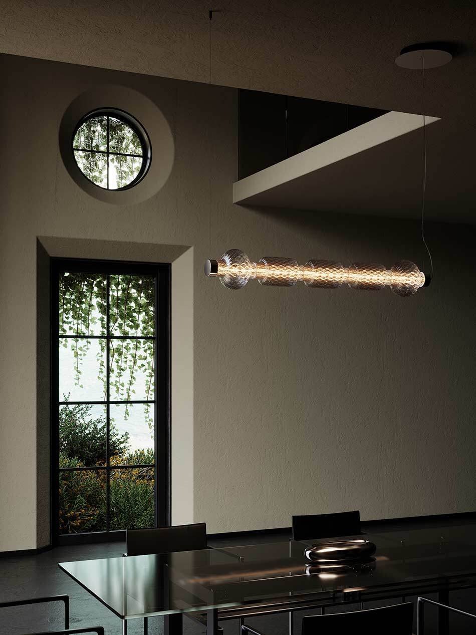 Modernes Interieur mit einer horizontalen Alambicco Pendelleuchte von Artemide aus mehreren transparenten Glaszylindern, die warmes Licht abstrahlen. Die Leuchte hängt über einem dunklen Esstisch. Im Hintergrund befinden sich ein hohes Fenster mit Blick ins Grüne sowie ein rundes Oberlicht. Das gedämpfte Licht betont die klare, architektonische Gestaltung des Raumes. Bild: Artemide - © Artemide