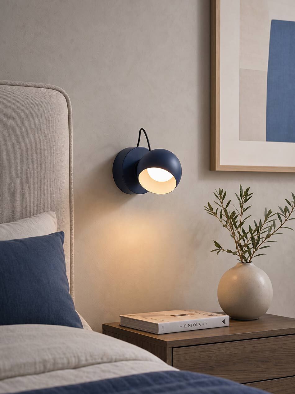 Modernes Schlafzimmer mit einer dunkelblauen Ixa Wandleuchte von Artemide über einem Nachttisch aus Holz. Die eingeschaltete Leuchte spendet warmes Licht. Auf dem Nachttisch stehen eine Vase mit Zweigen und ein Buch, darüber hängt ein gerahmtes Kunstbild. Links ist ein gepolstertes Bett mit blauen Kissen und blauer Bettwäsche zu sehen. Bild: Artemide – © Artemide | KI-generiert