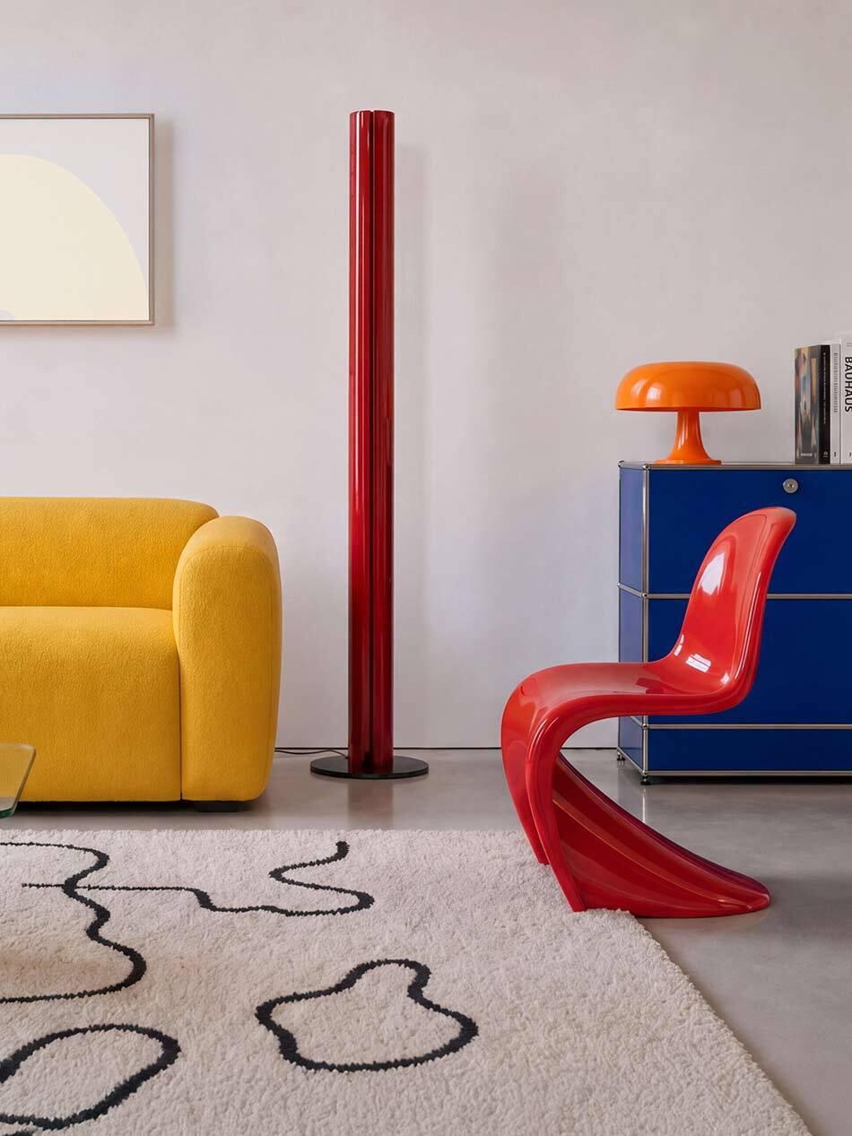 Farbenfroher Wohnraum mit einer roten Megaron Stehleuchte von Artemide, einem roten Freischwinger-Stuhl und einer orangefarbenen Nessino Tischleuchte von Artemide auf einem blauen Sideboard. Daneben steht ein gelbes Sofa. Ein heller Teppich mit abstraktem Muster ergänzt die moderne Einrichtung. Die kräftigen Farben der Möbel und Leuchten setzen markante Akzente im Raum. Bild: Artemide - © Artemide I KI-generiert
