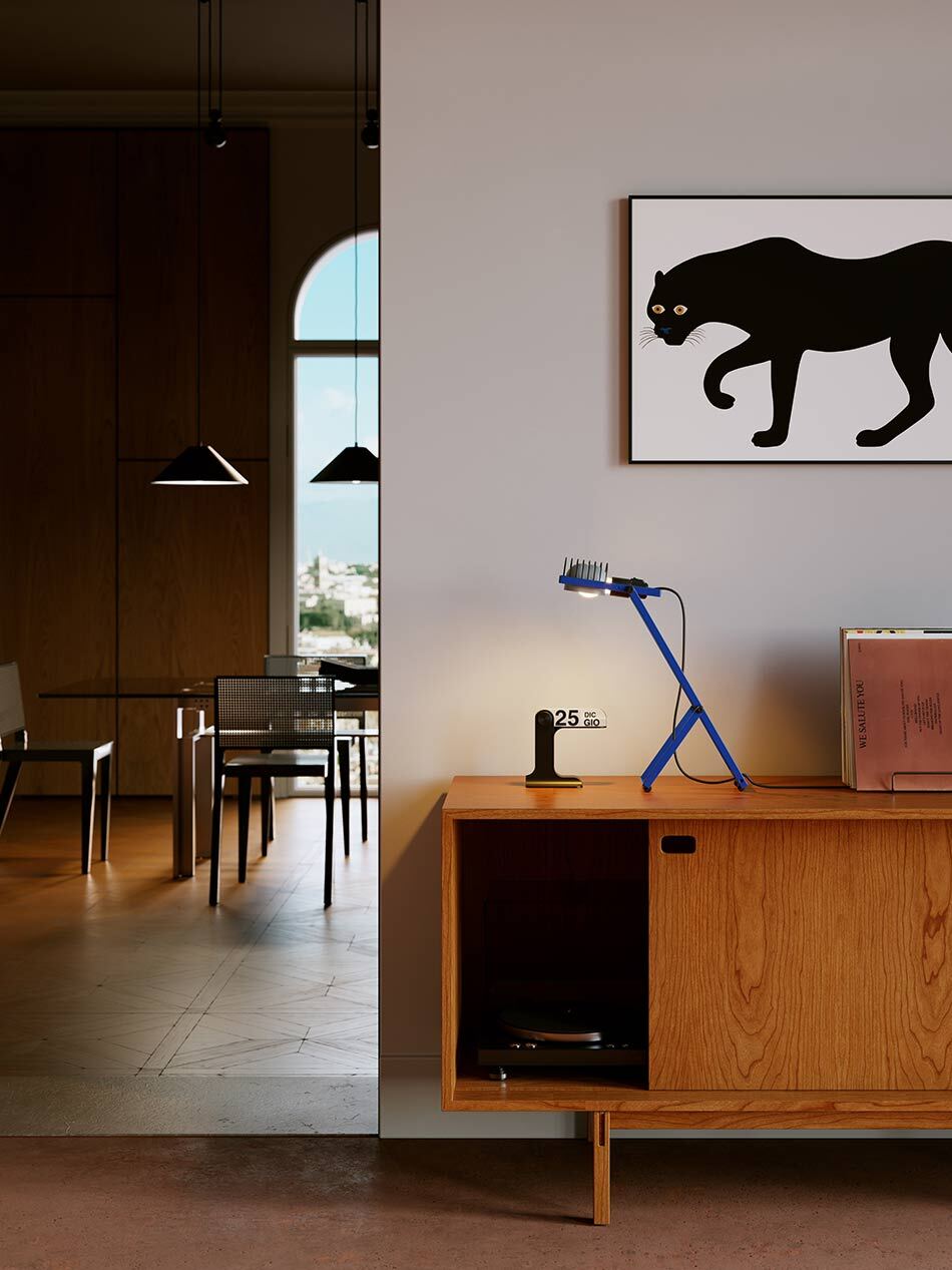 Wohnraum mit einem Sideboard aus Holz, auf dem eine blaue Sintesi Tischleuchte von Artemide steht und warmes Licht abstrahlt. Darüber hängt ein gerahmtes Bild mit einer schwarzen Katze auf hellem Hintergrund. Links öffnet sich der Raum zu einem Essbereich mit Tisch, Stühlen und schwarzen Pendelleuchten. Durch ein Rundbogenfenster fällt Tageslicht in den Innenraum. Bild: Artemide - © @b432visual @boccaccio432