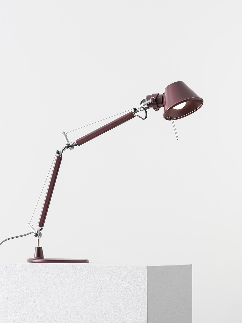 Verstellbare Tolomeo Micro Tischleuchte von Artemide in glänzendem Dunkelrot mit zwei gelenkig verbundenen Armen und konischem Leuchtkopf. Die Leuchte steht auf einem weißen Podest und ist durch sichtbare Zugfedern sowie Metallgelenke flexibel ausrichtbar. Das eingeschaltete Leuchtmittel spendet gerichtetes Licht. Die Kombination aus technischer Konstruktion und klarer Formensprache prägt das ikonische Design. Bild: Artemide - © Federico Villa