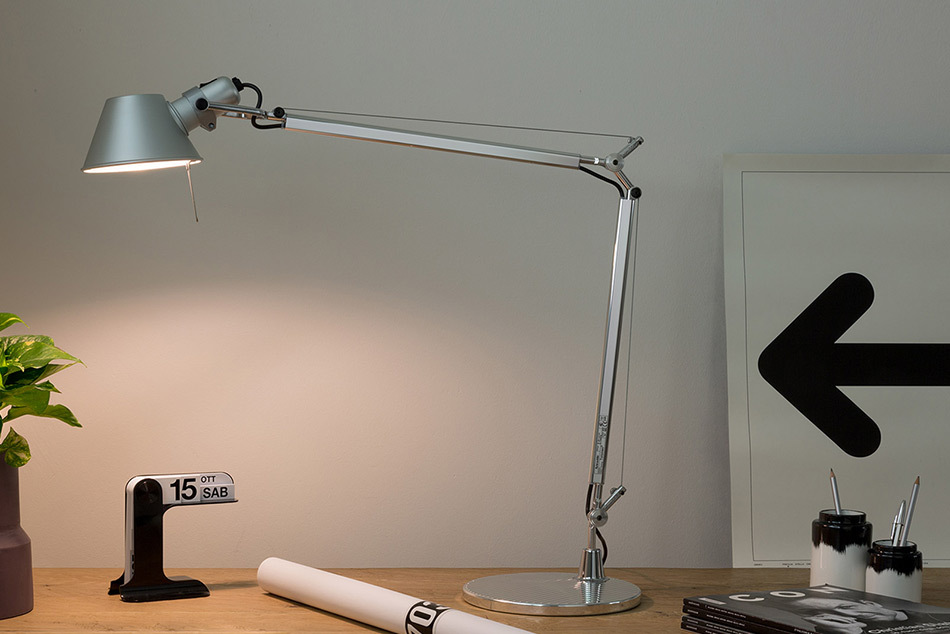 Silberfarbene Tolomeo Tavolo Schreibtischleuchte von Artemide mit verstellbarem Arm auf einem Holztisch. Die eingeschaltete Leuchte beleuchtet die helle Wand und die Tischoberfläche. Daneben stehen ein schwarzer Tischkalender, eine gerollte Zeichnung, Stiftehalter mit Stiften sowie ein gerahmtes Bild mit einem großen schwarzen Pfeil. Bild: Artemide – © Artemide