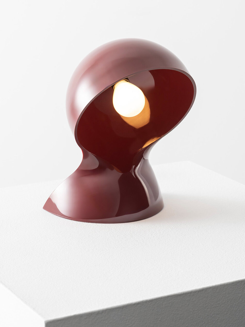 Skulpturale Dalu Tischleuchte von Artemide in glänzendem Dunkelrot auf einem weißen Podest. Der kuppelförmige Leuchtkopf ist nach vorne geneigt und gibt den Blick auf das eingeschaltete Leuchtmittel frei. Die organisch geschwungene Form und die monochrome Farbgestaltung setzen die Leuchte als Designobjekt in Szene. Bild: Artemide - © Federico Villa