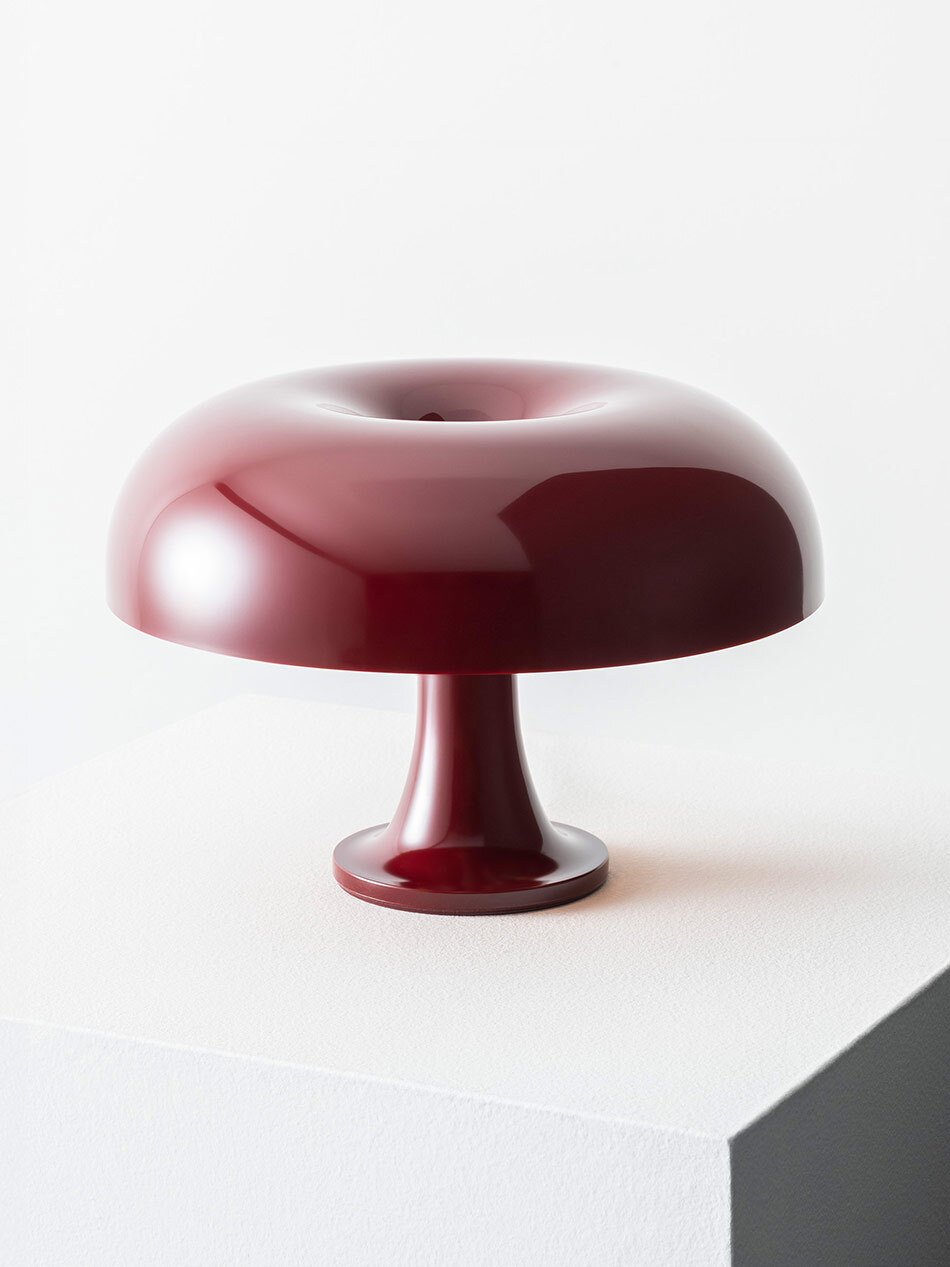 Pilzförmige Nessino Tischleuchte von Artemide in glänzendem Dunkelrot auf einem weißen Podest. Der breite, abgerundete Leuchtenschirm ruht auf einem schmalen, mittig platzierten Fuß. Die monochrome Farbgestaltung und die weichen, organischen Formen betonen den skulpturalen Charakter des Designobjekts. Bild: Artemide - © Federico Villa