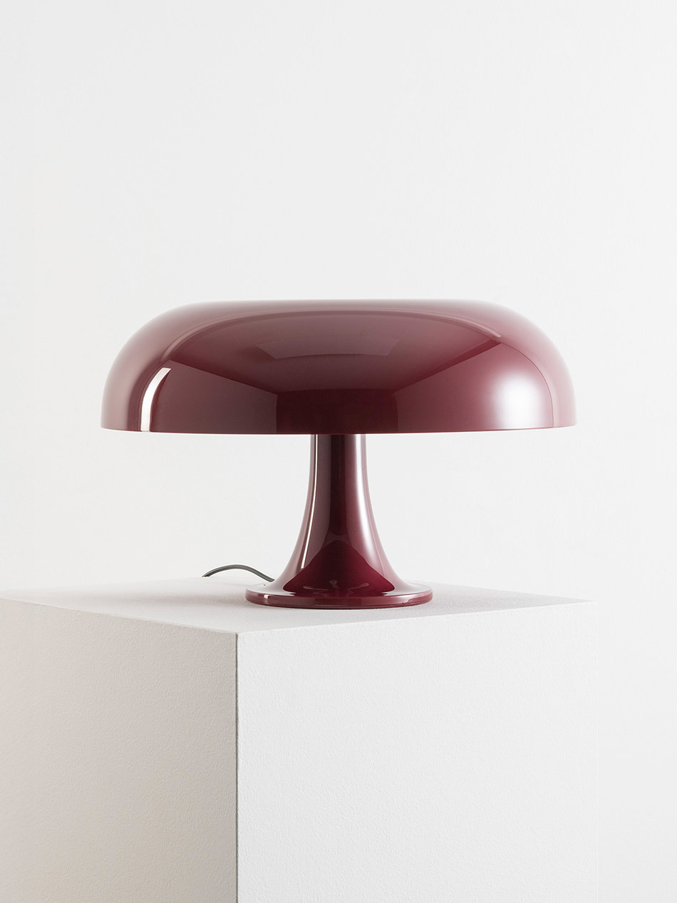 Pilzförmige Nesso Tischleuchte von Artemide in glänzendem Dunkelrot auf einem weißen Podest. Der breite, flach gewölbte Leuchtenschirm ruht auf einem schmalen, konisch geformten Fuß. Die monochrome Farbgestaltung und die klaren, weichen Linien betonen den skulpturalen Charakter der Leuchte als Designobjekt. Bild: Artemide - © Federico Villa