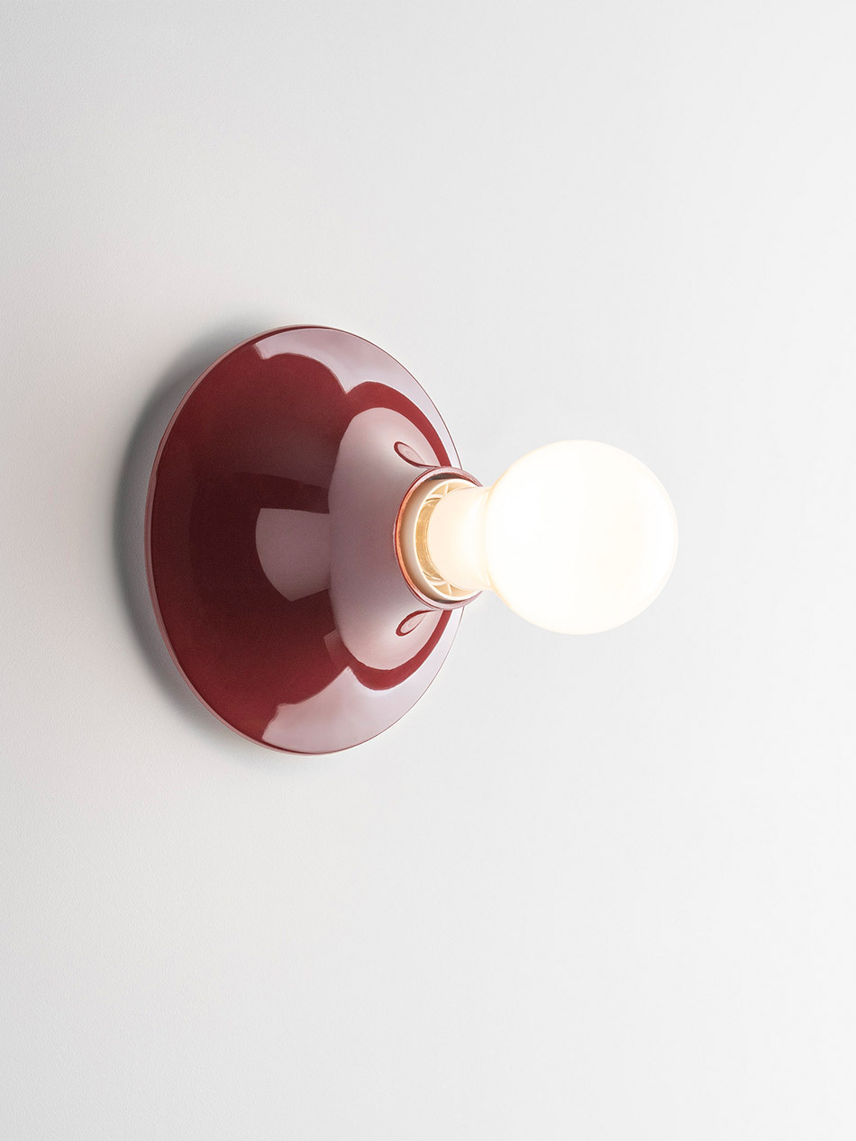 Teti Wandleuchte von Artemide in glänzendem Dunkelrot mit kreisförmiger Wandhalterung und freiliegendem kugelförmigem Leuchtmittel. Die Leuchte ist an einer hellen Wand montiert und strahlt gleichmäßiges, warmes Licht ab. Die einfache Formensprache hebt die Kombination aus farbiger Fassung und sichtbar eingesetztem Leuchtmittel hervor. Bild: Artemide - © Federico Villa