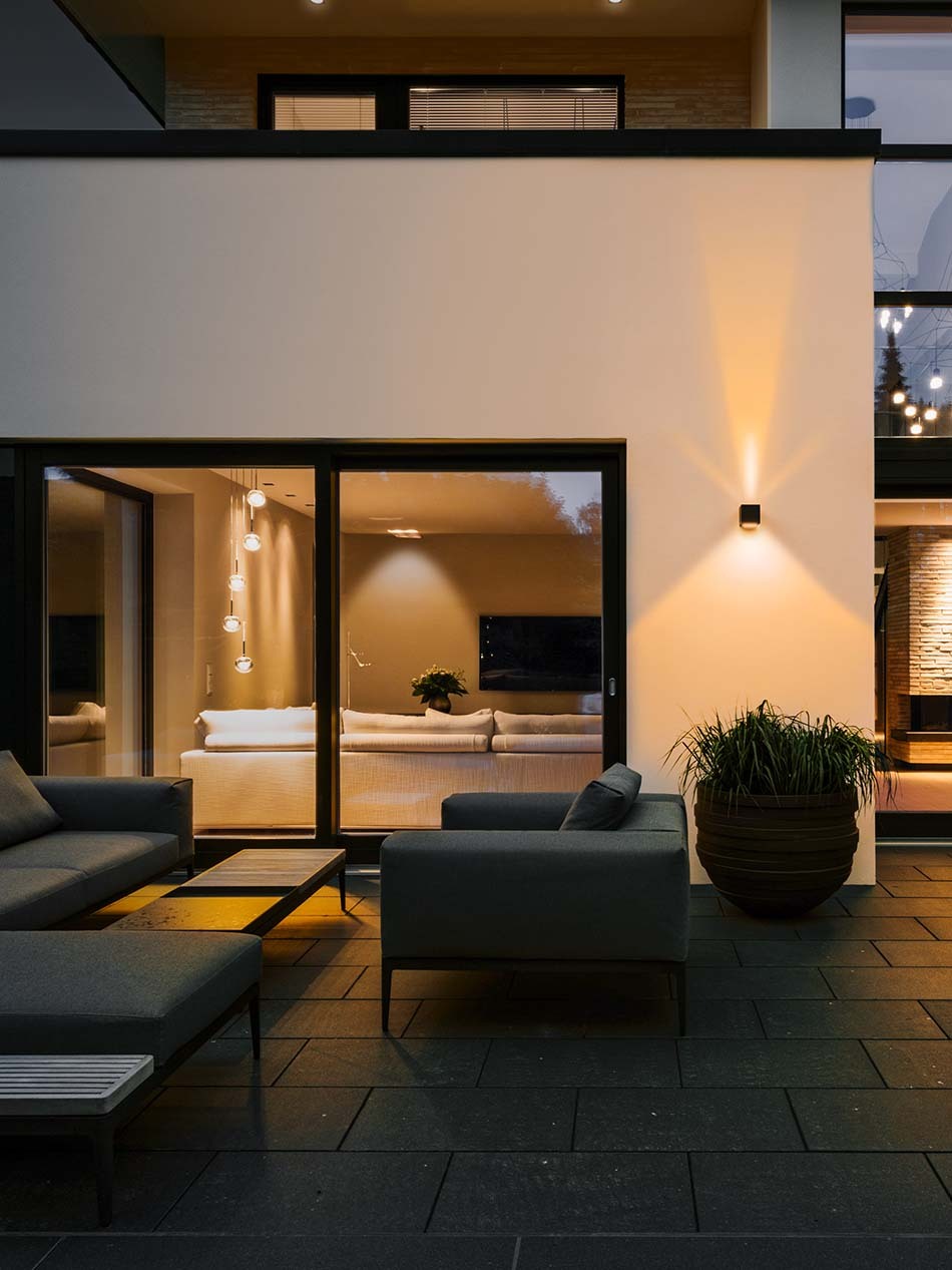 Terrasse mit schwarzer p.037 Wandleuchte von prediger.base mit zweiseitig strahlendem Licht. Auf der Terrasse befinden sich grauen Lounge-Möbel. Der Innenbereich wird durch die Dew Pendelleuchte von Kdln beleuchtet.
