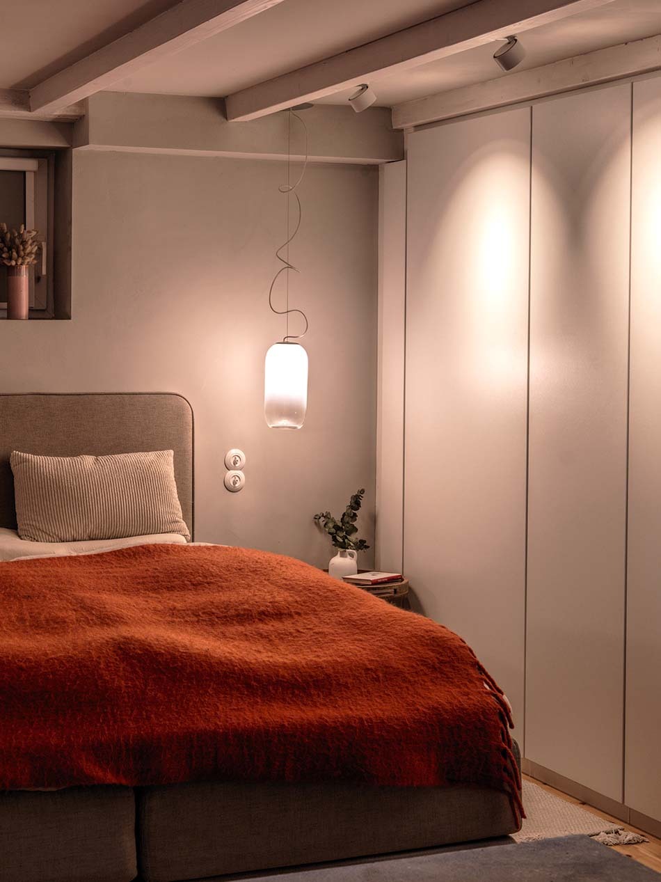 Schlafzimmer mit Gople Pendelleuchte von Artemide, die als Nachttischleuchte dient. Mehrere p.011 Deckenstrahler von prediger.base in weiß beleuchten den Kleiderschrank.