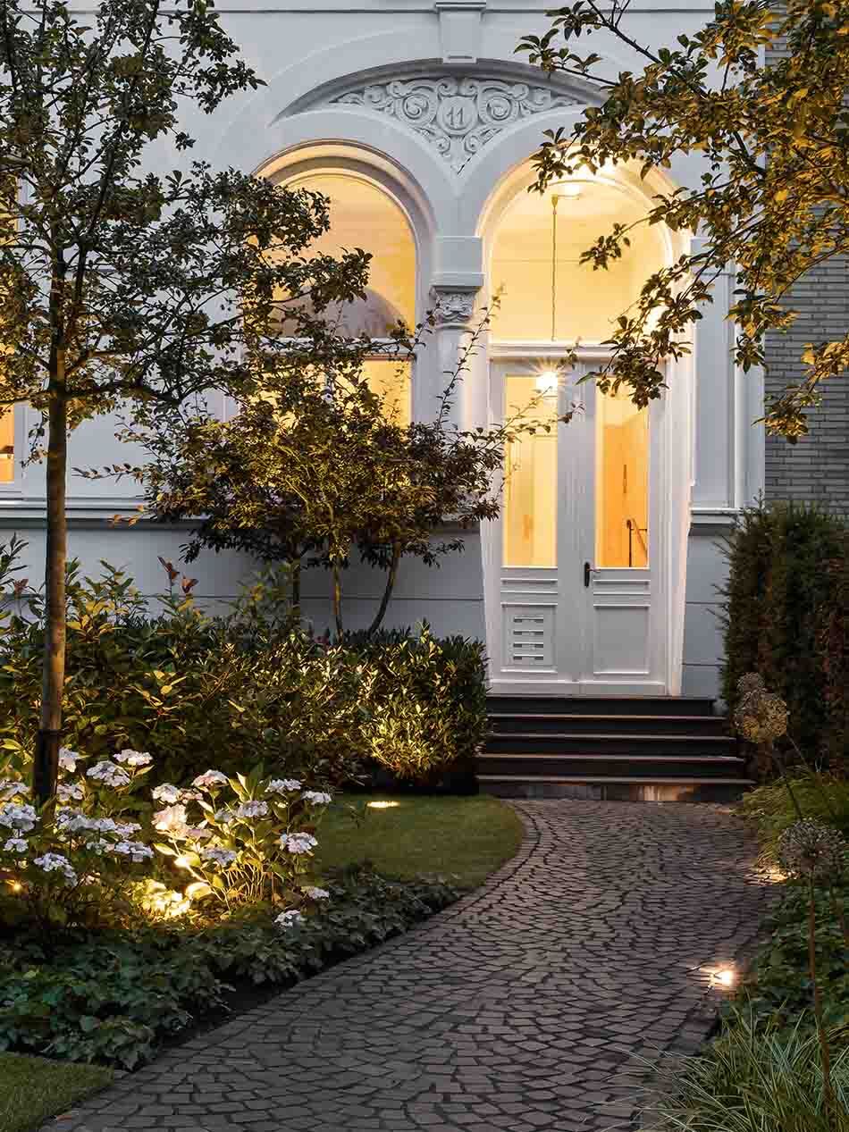 Garten mit mehreren Bodenstrahlern von Bega, die die vegetation sowie den gepflasterten Weg zur Eingangstür beleuchten. Es wurden unterschiedliche Pflanzen und Bäume gepflanzt.