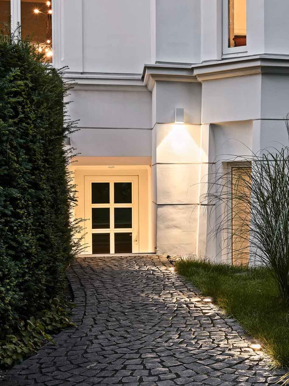 Fassade einer Altbauvilla mit einer weißen Wandleuchte von Bega mit einseitigem Lichtaustritt. Weitere Bodeneinbauleuchten von Bega beleuchten den gepflasterten Weg.