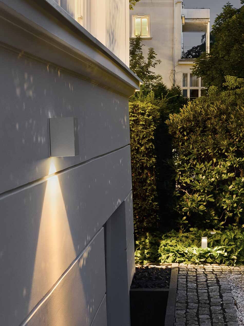 An einer Hausfassade befindet sich eine weiße Wandleuchte von Bega mit nach unten abstrahlendem Licht. Im Hintergrund ist der Garten zu sehen.