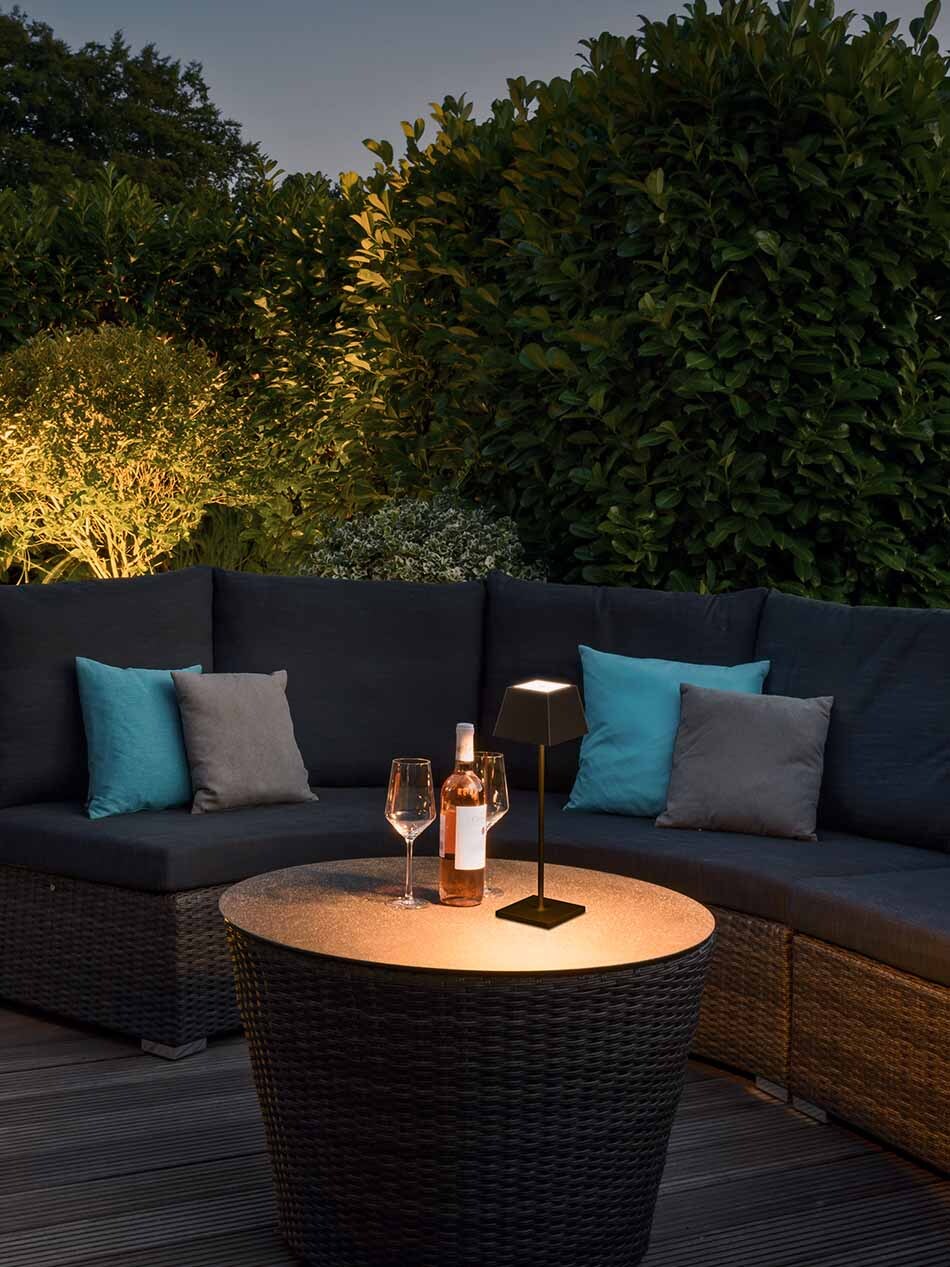 Terrasse mit Lounge-Bereich und einer schwarzen p.092 AkkuTischleuchte von prediger.base, die auf einem Tisch steht. Neben der Leuchte stehen Weingläser und eine Weinflasche.