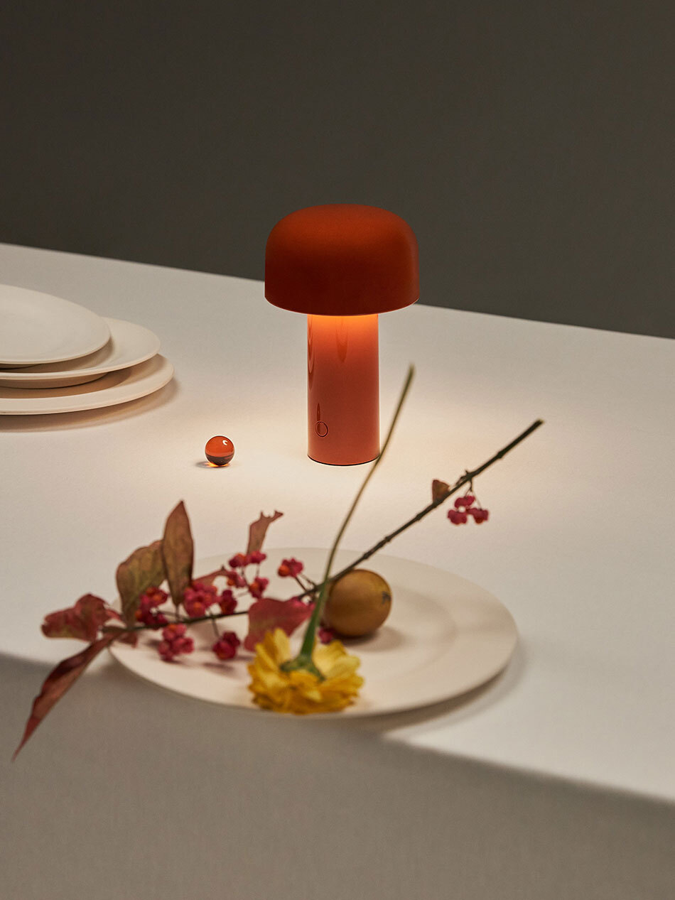 Ein gedeckter Esstisch mit weißer Tischleuchte, weißen Tellern und Deko-Blumen. In der Mitte steht die Bellhop Battery Akkuleuchte von Flos in Rot und leuchtet. (Bild: Flos)