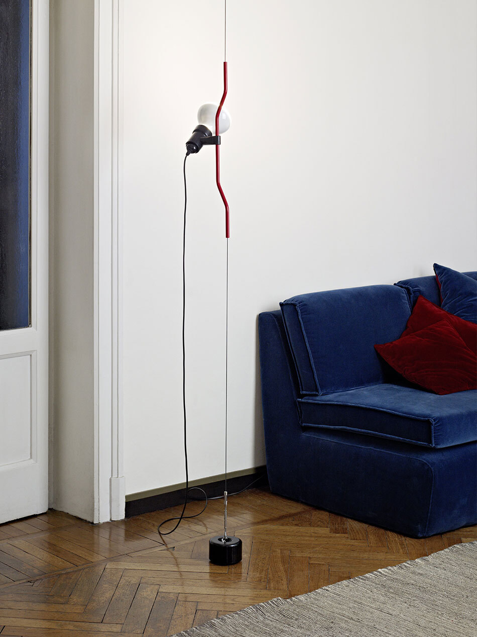Wohnzimmer mit Teppich und blauem Sofa mit rotem Kissen im Hintergrund. Im Vordergrund ist die Parentesi Leuchte von Flos in Rot zu sehen, die von der Decke hängt. (Bild: Flos)