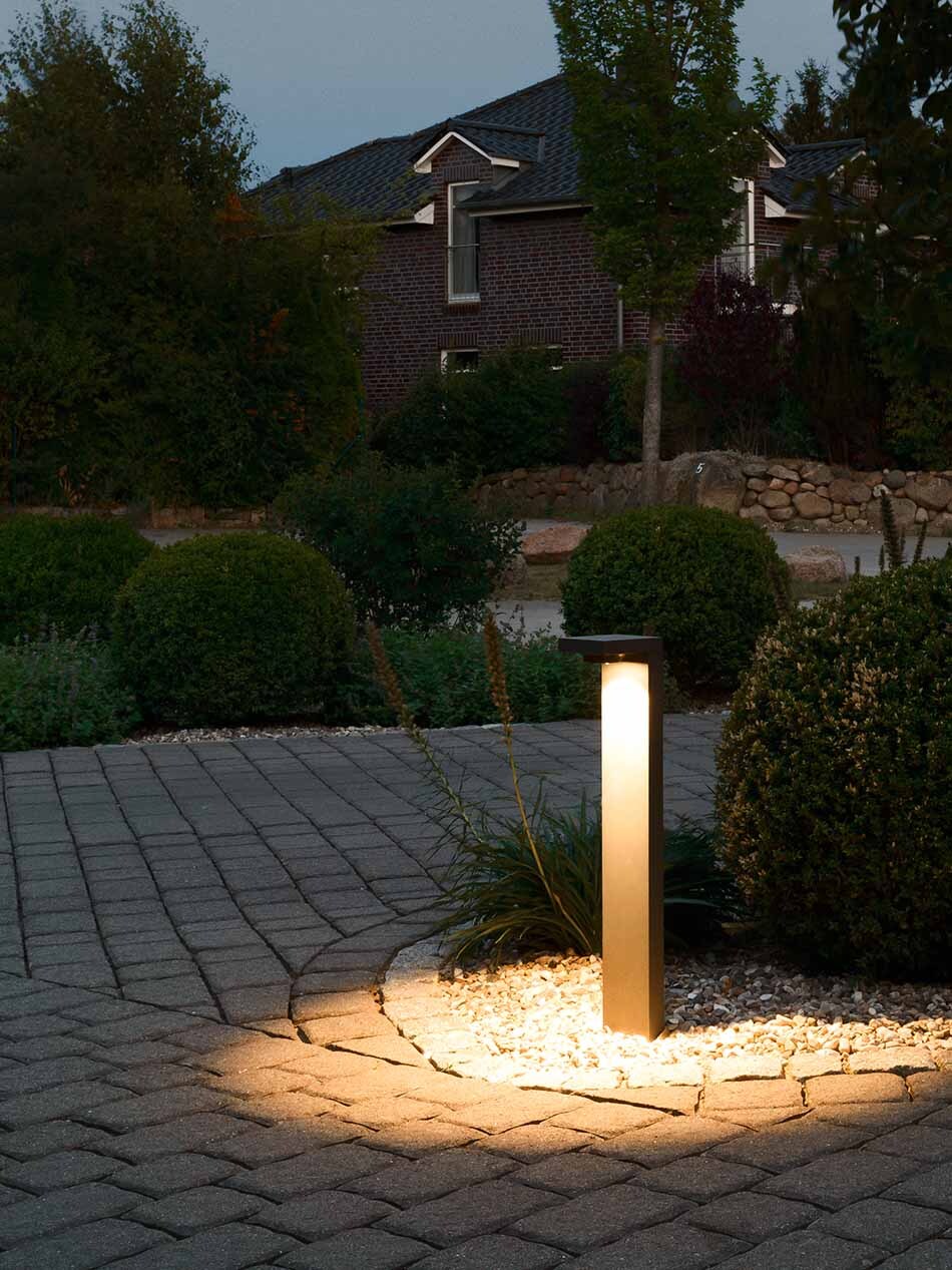 Einfahrt mit einer p.028 Pollerleuchte von prediger.base mit abgeblendetem Licht. Im Hintergrund sind unterschiedliche Büsche zu sehen.