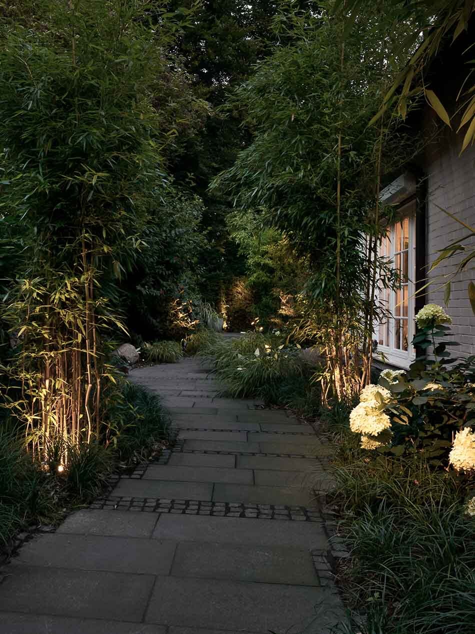 Großer Garten mit einem gepflastertem Weg und vielen unterschiedlichen Pflanzen, die mit mehreren p.119 Bodenstrahlern von prediger.base beleuchtet werden.