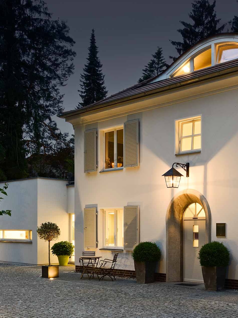 Das Bild zeigt ein großes Haus, das von meheren Lichtquellen beleuchtet wird. Über der Haustür hängt eine große schwarze Laterne. Neben dem Hauseingang stehen Sträucher, ein kleiner Tisch und zwei Stühlen. Überrollbare Boden-Scheinwerfern von Bega beleuchten die Fassade. Der Innenbereich des Hauses ist ebenfalls beleuchtet.