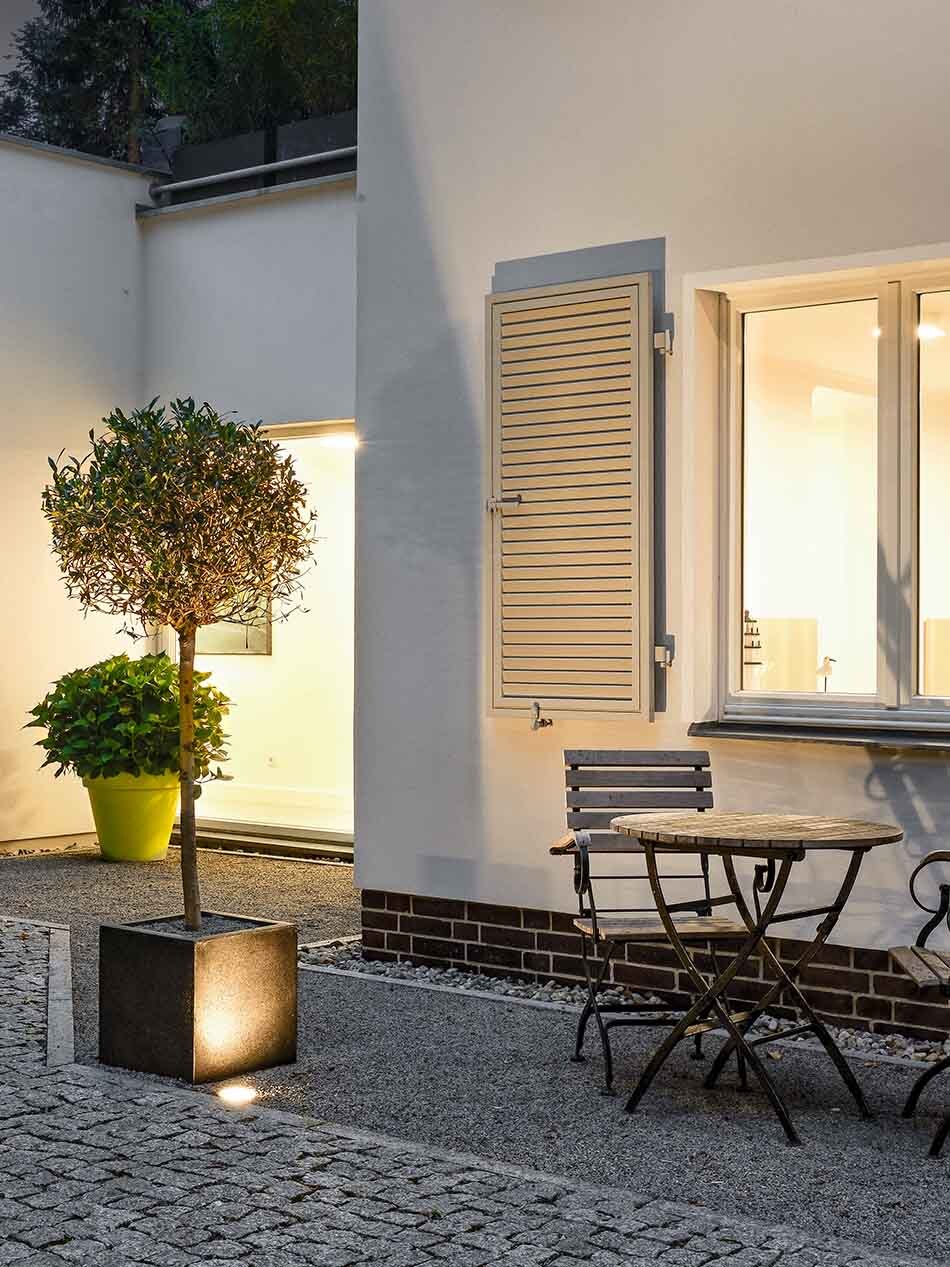 Terrasse mit Sträuchern, einem kleinen Tisch und zwei Stühlen sowie mit überrollbaren Scheinwerfern von Bega. Der Innenbereich des Hauses ist ebenfalls beleuchtet.