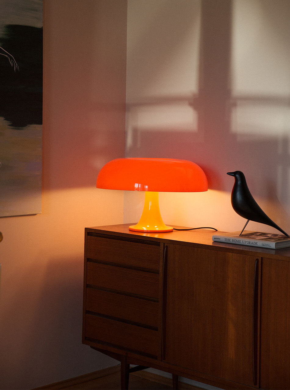 Auf einem Sideboard aus dunklem Holz steht eine Nessino Tischleuchte in Orange sowie ein Dekovogel. Die Leuchte verströmt warmes Licht.