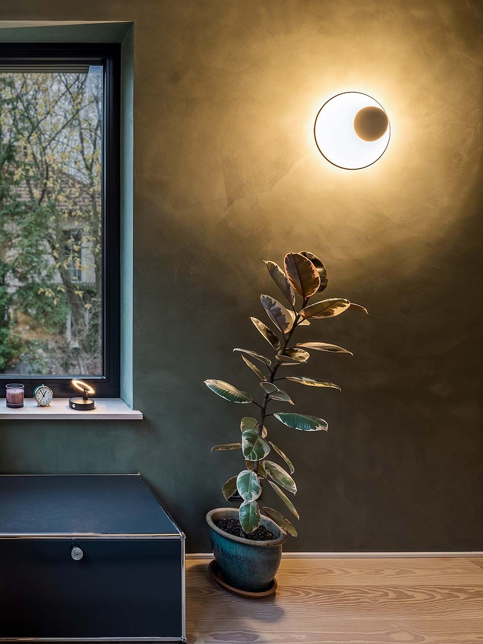 Modernes Wohnzimmer mit einer Funnel Wandleuchte von Vibia, die die dunkelgrüne Wand beleuchtet.