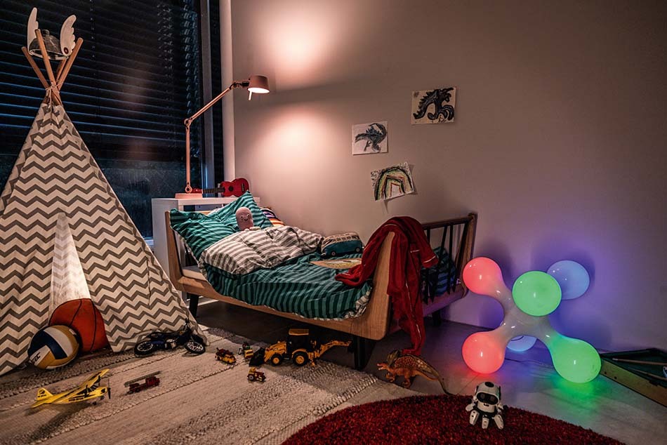 Ein buntes Kinderzimmer am Abend mit einer Bolt Tischleuchte von Tonone in Rosa, die über dem Bett steht. Vor dem Bett steht eine Atomium Bodenleuchte von KDLN mit bunten Leuchtmitteln. Links neben dem Bett steht ein Spielzelt mit kleinen Spielfiguren. An der Wand hängen selbst gemalte Bilder.