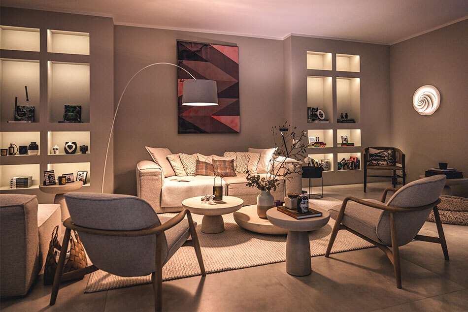 Modernes Wohnzimmer mit unterschiedlichen Lichtquellen. Neben dem Sofa steht eine beige Twiggy Bogenleuchte von Foscarini. An der Wand hängt die Wandleuchte Swirl von Le Klint. In den Einbauschränke befinden sich prediger.base Einbauleuchten.