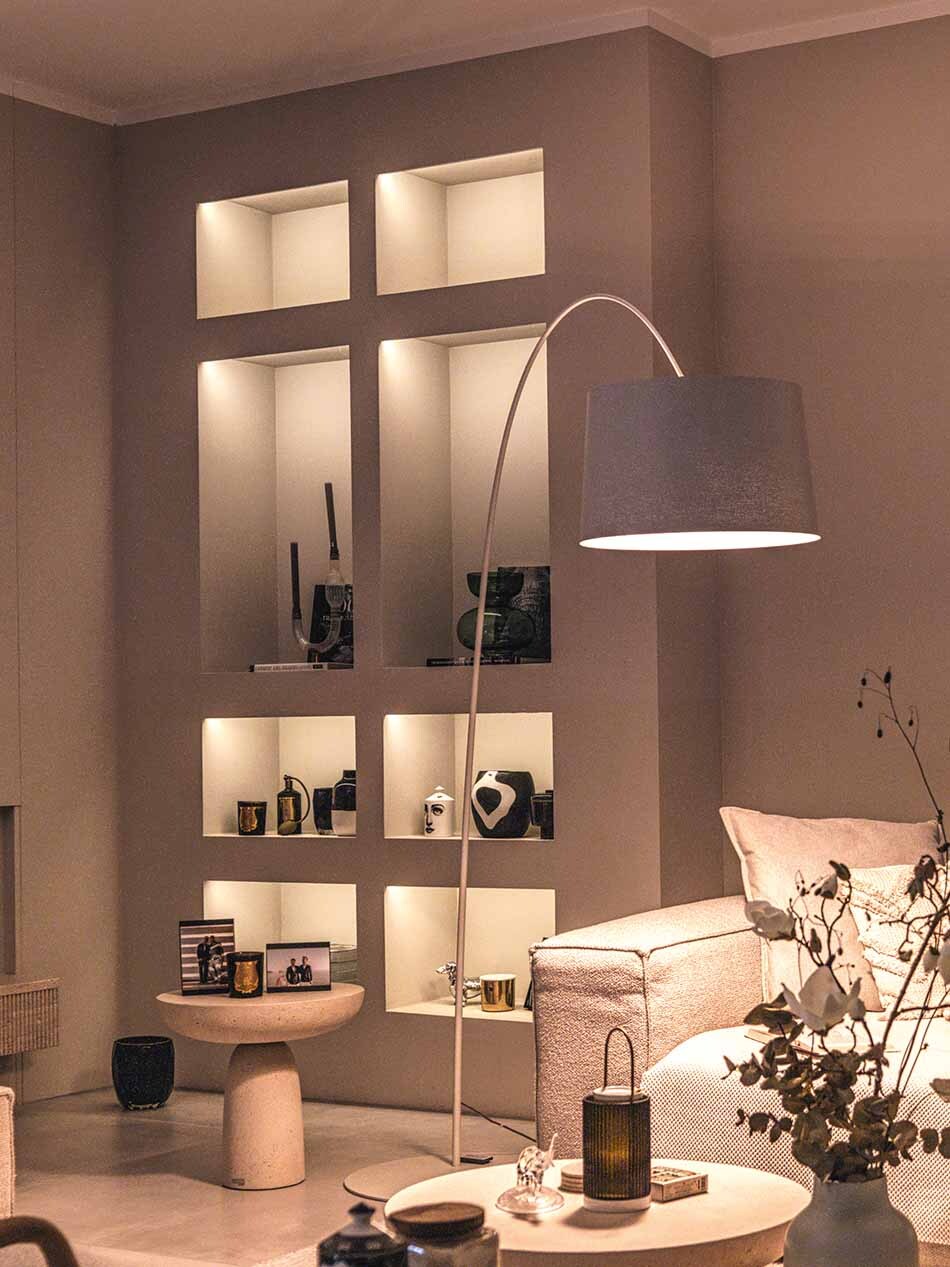 Modernes Wohnzimmer einer Villa mit unterschiedlichen Lichtquellen. Neben dem Sofa steht eine Bogenleuchte Twiggy von Foscarini. Die Wandregale werden von Lichtleisten von prediger.base beleuchtet. Das Wohnzimmer ist gemütlich eingerichtet.