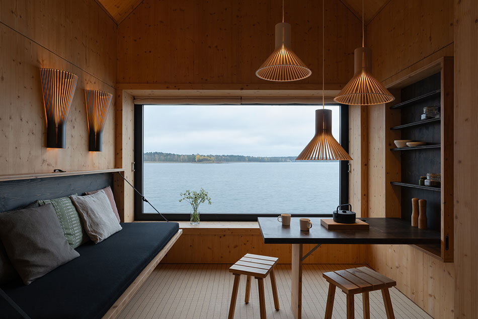 Warm beleuchteter Holzraum mit mehreren Leuchten von Secto Design. Pendel- und Wandleuchten aus Holz erzeugen ein gemütliches Lichtambiente. Im Hintergrund bietet ein großes Fenster Blick auf einen See.