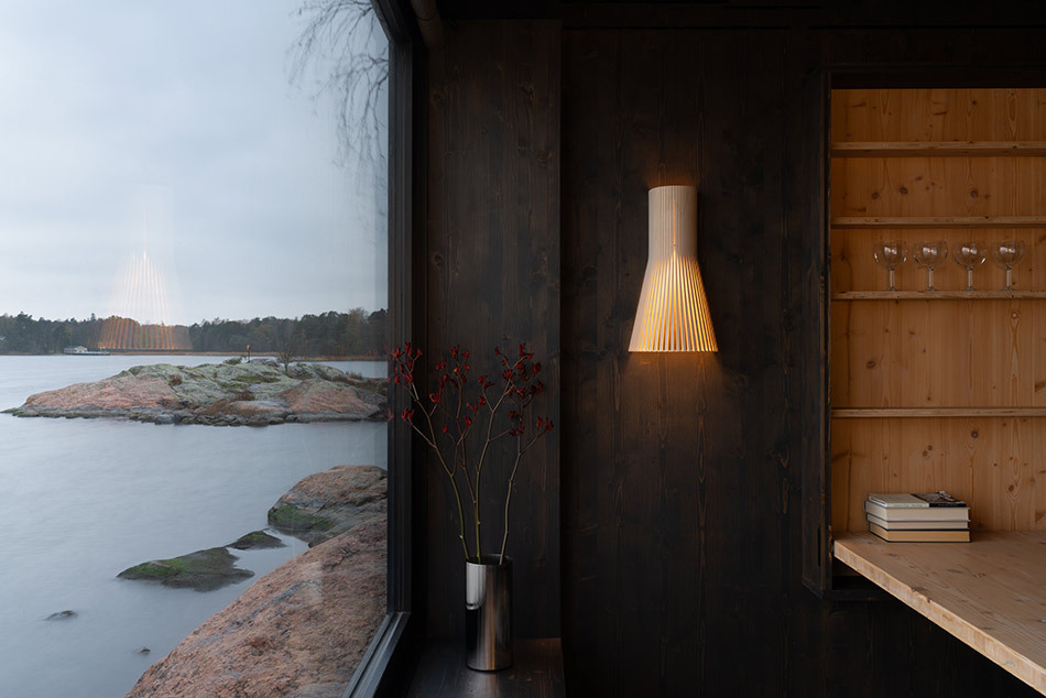 Secto Design Wandleuchte Secto 4231 in einem modernen Holzinterieur. Die Leuchte spendet warmes Licht und ist an einer dunkel gebeizten Holzwand montiert. Daneben befindet sich ein großes Fenster mit Blick auf eine felsige Küstenlandschaft am Wasser. Rechts sind ein offenes Regal mit Gläsern und Büchern sowie ein kleiner Blumenstrauß in einer Vase zu sehen.