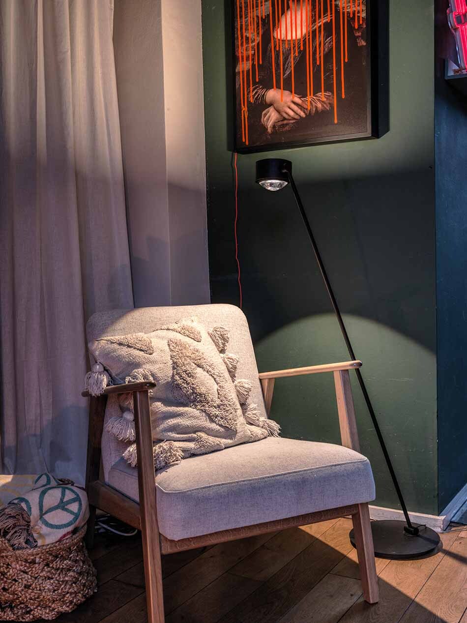 Das Bild zeigt eine Ecke eines Wohnzimmers, in der ein Lounge Stuhl und eine s.200 Stehleuchte in satin schwarz von prediger.select steht. Die Leuchte beleuchtet den Stuhl sowie die dunkelgrüne Wand mit Kunstwerken.