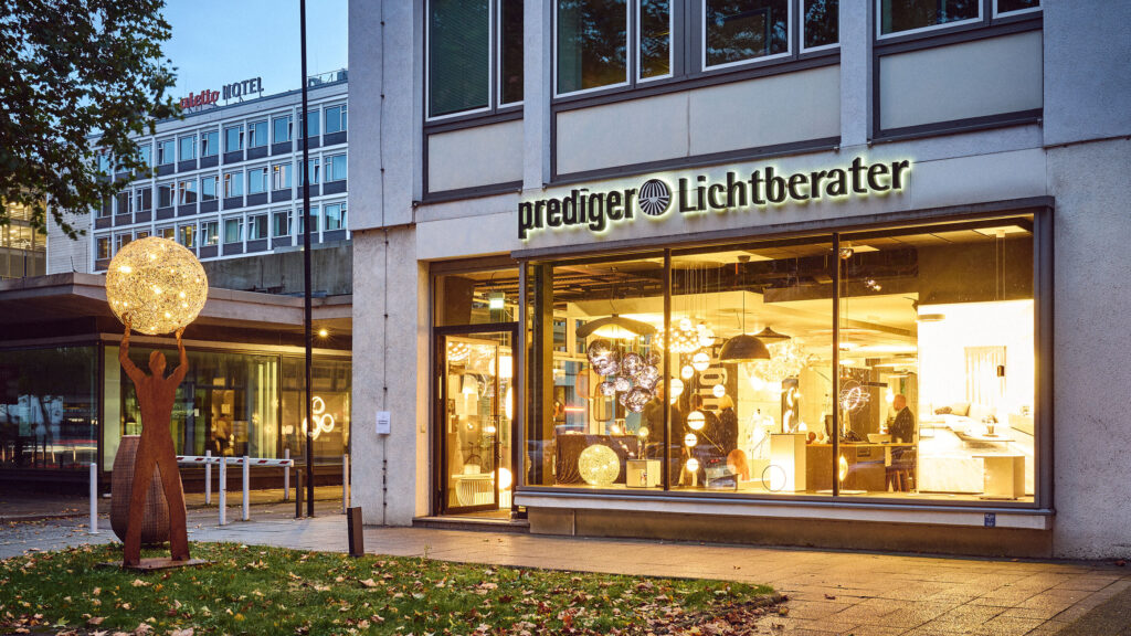 Das Bild zeigt den Showroom von Prediger Lichtberater in Berlin bei Tageslicht. Die große Glasfassade ermöglicht einen klaren Blick in den stilvoll eingerichteten Innenraum, in dem eine Vielzahl von modernen Leuchten ausgestellt sind. Vor dem Eingang steht eine Skulptur aus Metall, die eine Figur darstellt, die eine große, leuchtende Kugel in den Händen hält.